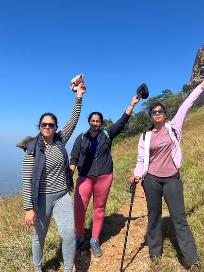 Three trekkers celebrating on the trail, with the Kurinjal peak and blue sky behind them.
