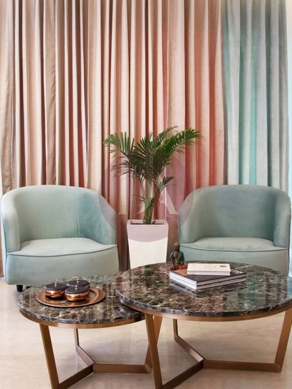 This cozy corner features two mint-green armchairs and nested marble coffee tables. The dual-toned pink and green curtains add a soft, layered backdrop, demonstrating our focus on texture and subtle color play.