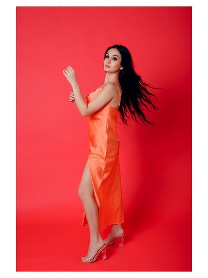 A vibrant, full-length fashion shot against a solid red background. The color contrast makes the orange dress pop, creating a bold and eye-catching image for a lookbook or campaign.