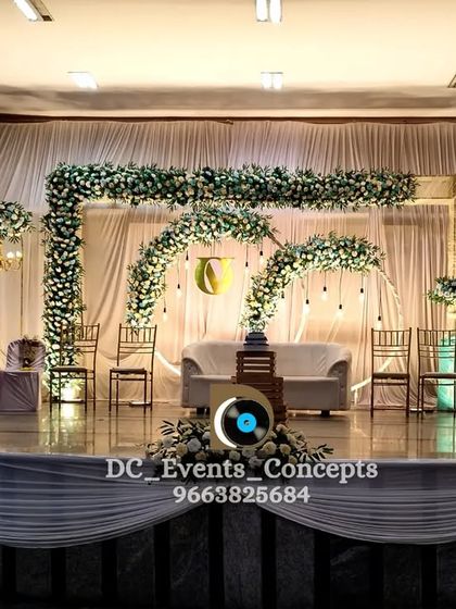 A fresh and airy wedding reception stage with white and green florals forming multiple arches. The design uses simple drop lights and clean white drapery for a minimalist yet elegant look.