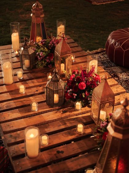 A close-up of the Moroccan lanterns and candles on the wooden pallet table. The mix of different lantern shapes and sizes, combined with the scattered candles, creates a beautiful, glowing centerpiece.