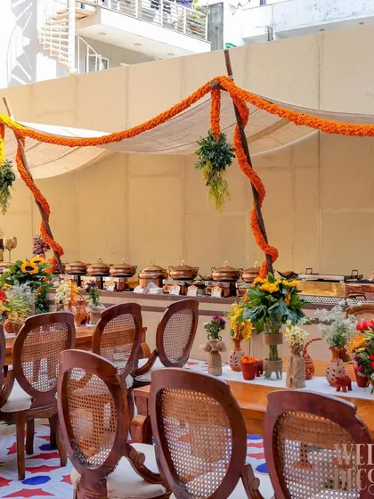 The dining and buffet setup under a simple canopy draped with thick marigold garlands. The wooden chairs and earthy tones were chosen to complement the rustic, traditional theme of the party.