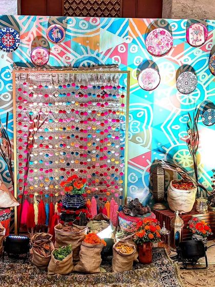 A creative and textural photo booth inspired by a spice market. The backdrop features a colorful patterned wall, a curtain of sequins and tassels, and bags of spices and flowers, creating a multi-sensory experience.
