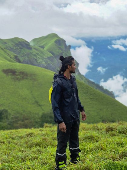A trekker in a raincoat looks out at the dramatic sky and green hills of Bandaje.