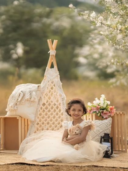 A girl in a beautiful dress sits with her teddy bear in a teepee setup in a field of white flowers, creating a whimsical outdoor portrait.