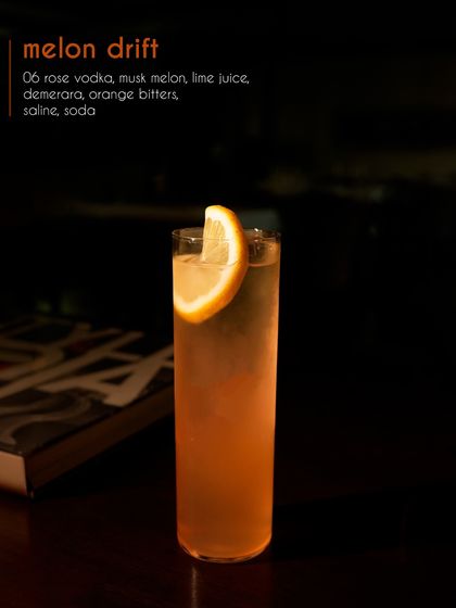 The Melon Drift, presented in a tall glass, glows with a gentle orange hue. This vodka-based highball combines musk melon and lime for a refreshing and subtly sweet flavor, perfect for a warm evening.
