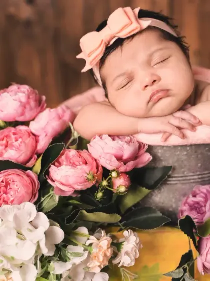 A beautiful newborn girl rests in a bucket surrounded by a bouquet of pink and purple flowers. A calendar block marks her birth date, making it a perfect birth announcement photo.