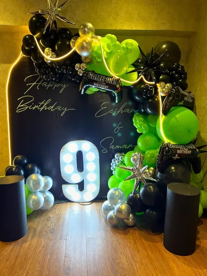 Get your game on with this epic gaming-themed ninth birthday party decor, featuring a bold black backdrop with neon-style light outlines and personalized "Happy Birthday" text. The setup is energized by a large light-up number '9' and a dynamic balloon arch in electric green, black, and silver, complete with gaming controller foil balloons.