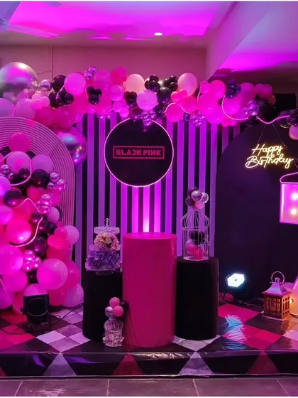 A trendy Blackpink themed birthday party, using a dramatic pink and black color scheme with a striped backdrop, balloon garlands, and a large light-up number '7'.