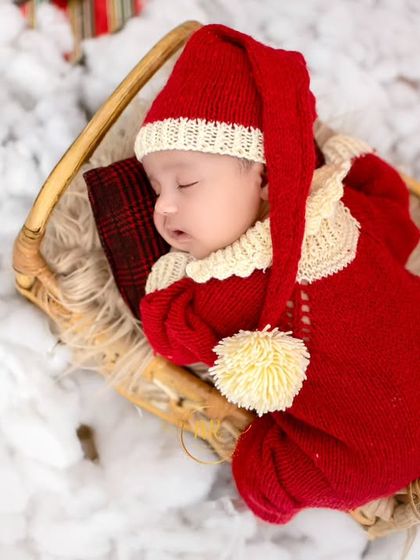 An overhead shot of a sleeping newborn in a red knit outfit and long Santa hat, nestled in a basket on a bed of fluffy white "snow".