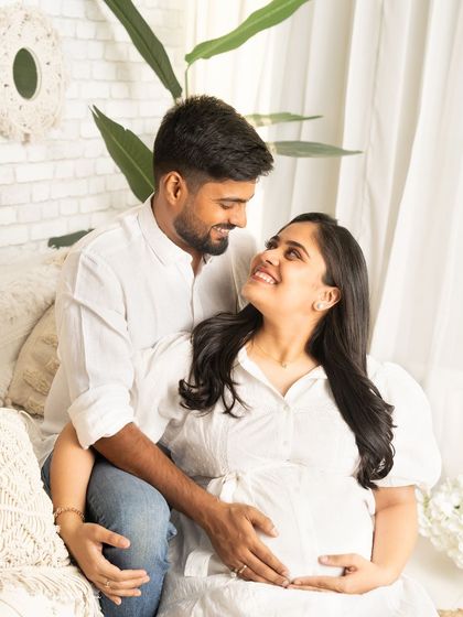 A happy and relaxed couple's portrait. Seated together in a bright, white-themed room, they look at each other with love and excitement for their new journey.