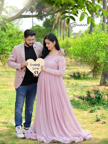 A lovely couple's portrait in a garden, holding a "Coming Soon" sign. The mother-to-be is wearing a pink lace gown.