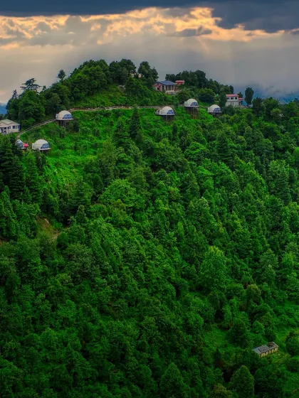 A wide shot of the Kanatal dome stay, capturing the property under dramatic sun rays breaking through the clouds. This image highlights the magical, dream-like quality of a glamping adventure.