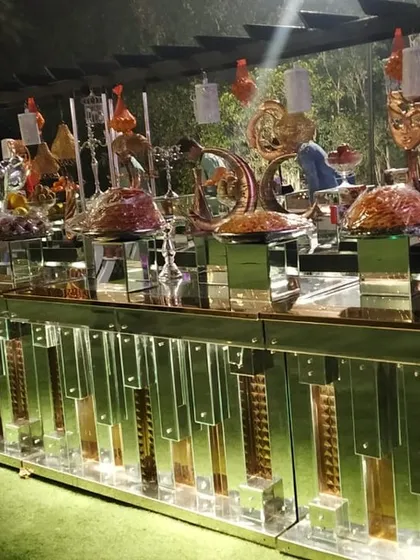 An expansive buffet setup on a lush green lawn for an outdoor event. The long, mirrored counter reflects the ambient light, creating a beautiful effect in the evening.