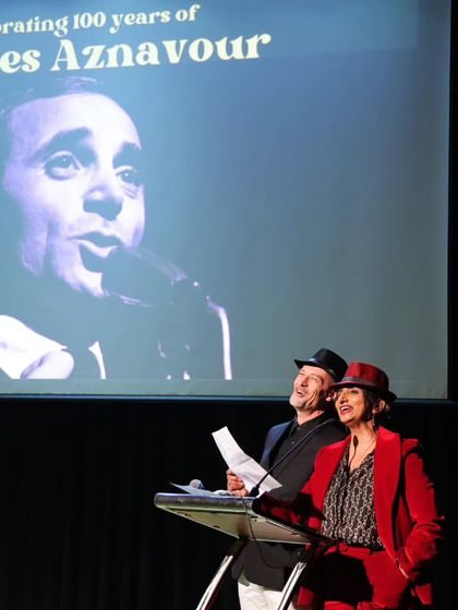 The hosts of the Aznavour tribute on stage, with a projection of the iconic singer behind them. The presentation is both a tribute and a celebration of his legacy.