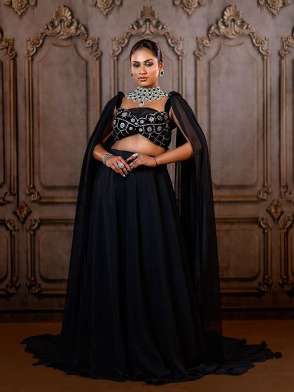 A powerful and elegant pose in a black lehenga with a cape. The ornate backdrop provides a beautiful contrast to the modern silhouette of the outfit.