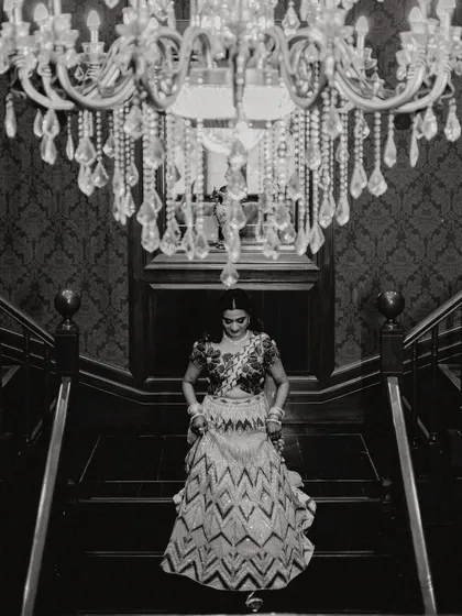 A beautiful black and white shot of the bride descending the stairs, framed by a crystal chandelier. This is an example of our artistic, cinematic approach to bridal portraits.