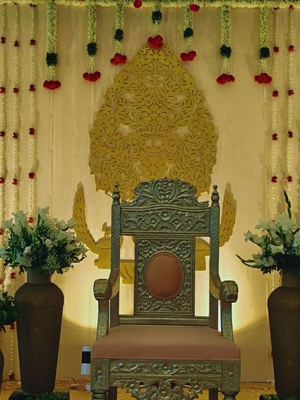 An elegant decor piece featuring a traditional throne against a dark green backdrop, accented with golden palm leaves and strings of jasmine flowers.