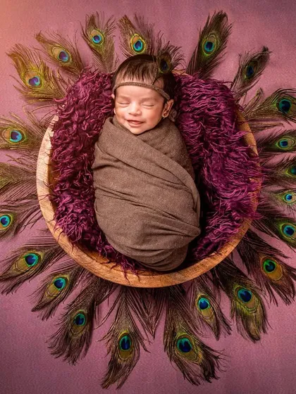 An artistic and elegant portrait, this overhead shot shows a newborn swaddled peacefully in a wooden bowl, encircled by a stunning halo of peacock feathers. The rich colors and textures create a breathtaking piece of art.
