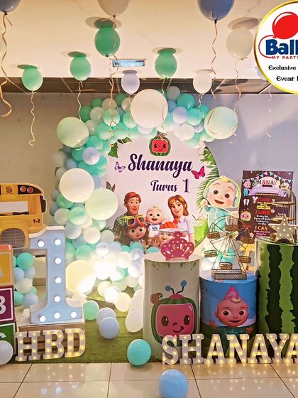 A full-stage Cocomelon-themed first birthday. The decor includes a pastel balloon arch, the iconic yellow bus, and a 'SHANAYA' marquee.