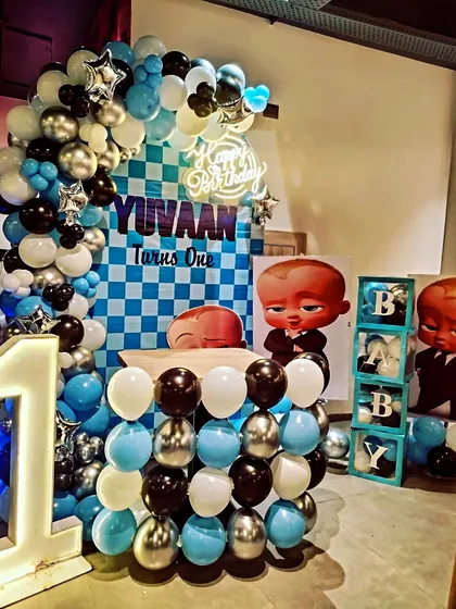 A fun and popular Boss Baby theme for a first birthday. This setup includes a checkerboard backdrop, character cutouts, "BABY" blocks, and a large light-up number "1", all in a classic blue, black, and silver color scheme.