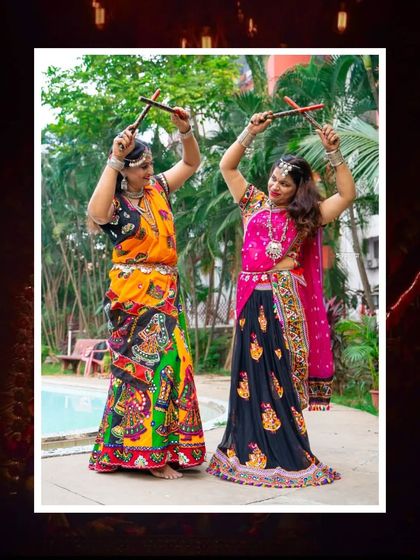 Two women strike a pose with dandiya sticks during a Garba photoshoot. Their colorful traditional outfits and joyful expressions perfectly capture the high energy and celebratory mood of Navratri.