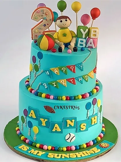 A colorful two-tier cake for a second birthday. This playful design features a fondant boy, a ball, alphabet blocks, and lots of balloon decorations.