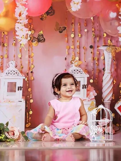 A ten-month-old baby girl smiling in a pink and gold-themed setup with balloons and lanterns.