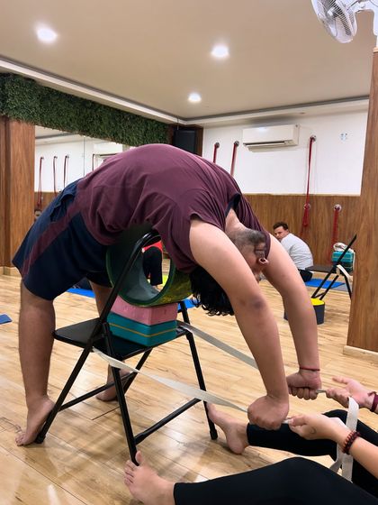 Teamwork in action. A partner provides gentle resistance with a strap while the student uses a chair and wheel to deepen their backbend, ensuring a safe and effective stretch.