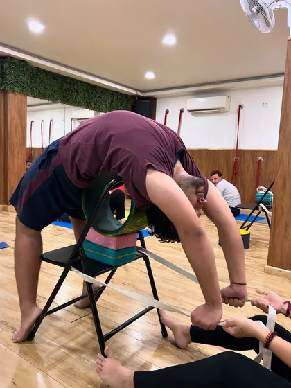 Teamwork in action. A partner provides gentle resistance with a strap while the student uses a chair and wheel to deepen their backbend, ensuring a safe and effective stretch.