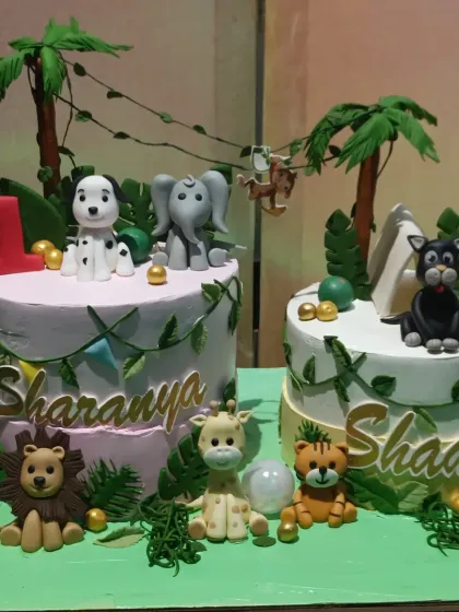 A pair of adorable jungle-themed cakes for a twin's first birthday. Each cake is personalized with different fondant animals like a dalmatian, elephant, lion, and tiger.