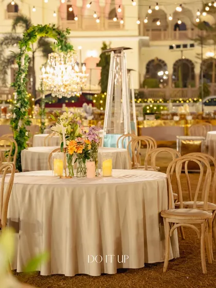 A wider view of the outdoor dinner seating in Jaipur. The round tables are elegantly set with simple floral centerpieces and candles, under a canopy of string lights.