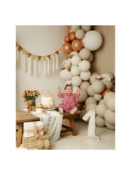 One year old and ready to celebrate. This little girl looks adorable in her birthday dress, sitting proudly next to her cake in a boho-themed studio setup with a balloon arch.