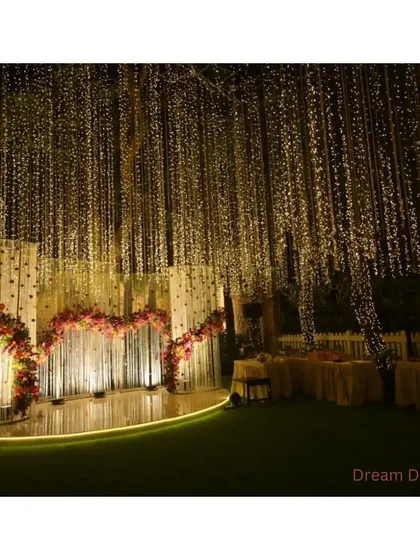 A dramatic stage setup for an evening event. A curtain of fairy lights provides a sparkling backdrop for a curved stage adorned with deep red floral arrangements.