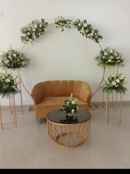 A minimalist and modern indoor setup for a small ring ceremony or engagement. It features a simple golden circular frame with white floral arrangements, a stylish sofa, and a matching coffee table.