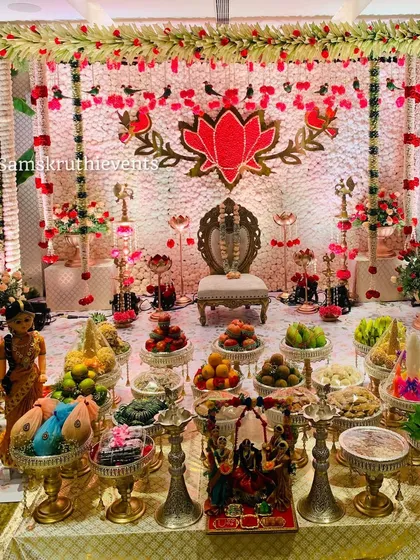 A full view of a puberty ceremony setup. The stage is filled with a lavish display of offerings, traditional dolls, and brass lamps, all set against a backdrop with a central lotus theme.