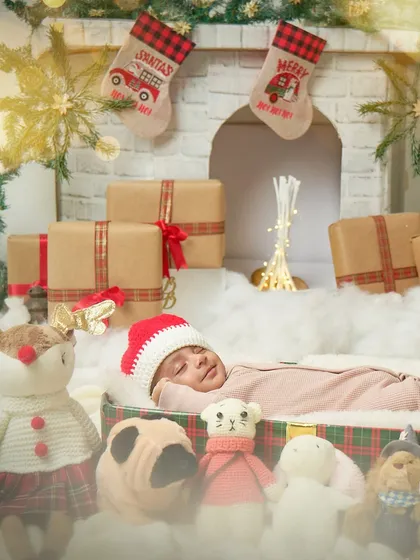 A wide view of the newborn Christmas setup, with the baby sleeping peacefully in a gift box surrounded by festive toys and a warm fireplace.