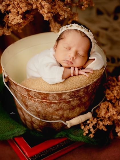 A rustic and warm newborn portrait. The baby sleeps peacefully in a metal bucket, with dried flowers adding a touch of natural, earthy texture.