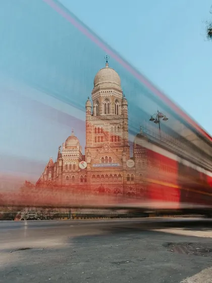 A long-exposure shot of a red BEST bus speeding past the BMC building, creating a beautiful motion blur that contrasts with the static heritage structure.