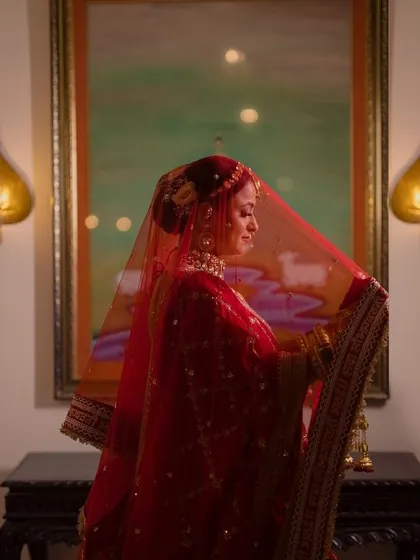 A moody and romantic bridal portrait, with the bride standing under her red veil in a softly lit room, creating a feeling of quiet contemplation.