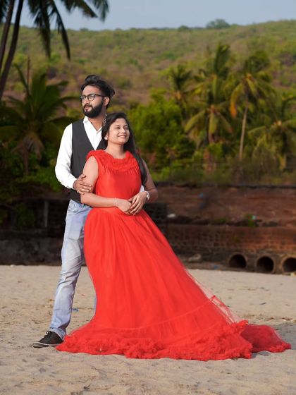 A relaxed pre-wedding photo on the beach. The bride-to-be is seated gracefully in her red ruffled gown, posing with her partner.