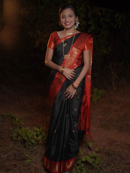 A full-length portrait showcasing the elegance of the traditional black saree worn during Makar Sankrant. Her gentle smile and poised stance against the dark, natural background make the colors of her attire pop.