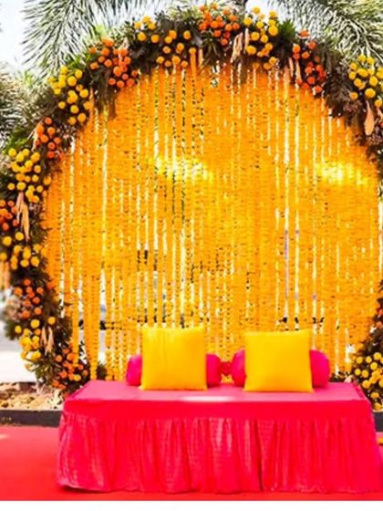 A vibrant outdoor Haldi photo booth with a circular backdrop of marigold strings. The bright pink seating provides a beautiful pop of color against the yellow flowers.