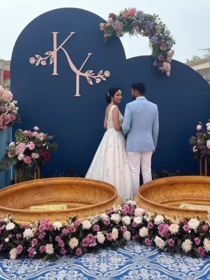 A personalized photo-op area for a Haldi or engagement function. This setup features a deep blue backdrop with the couple's monogram, framed by beautiful pink and lilac floral arrangements.