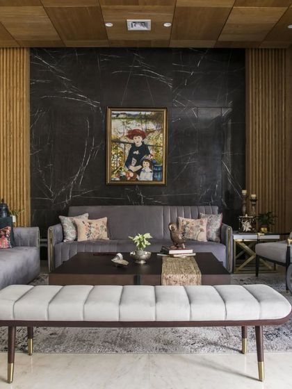 This living room combines a dramatic black marble accent wall with warm wood paneling. The plush, grey-toned furniture and a tufted bench create a comfortable yet sophisticated seating area, grounded by a patterned rug.