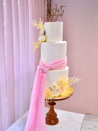 A dreamy and elegant three-tier engagement cake. Two tiers of blueberry and one of caramel are wrapped in pristine white fondant, accented with a flowing pink sash and delicate sugar flowers for a soft, romantic look.