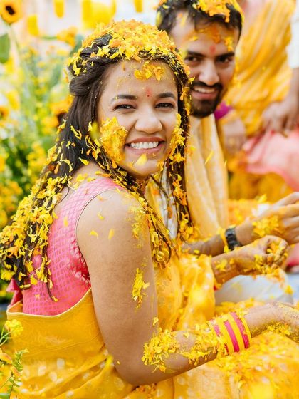 A radiant portrait of the bride, her face covered in turmeric and flowers, smiling brightly for the camera.