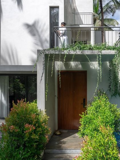 The east entrance to the SANCTUM House, nestled amongst lush bushes and climbing plants. The design creates a welcoming and private entry sequence, with a balcony overhead providing shade and a sense of enclosure.