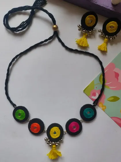 A playful and colorful handmade necklace set. This design features thread-wrapped circles in multiple colors on a black cord, with matching earrings that have bright yellow tassels.