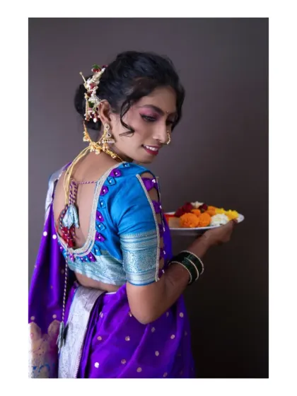 A classic Maharashtrian portrait. The model is dressed in a traditional purple saree with festive jewelry, holding a plate of flowers. The over-the-shoulder pose is a timeless way to showcase the intricate blouse design and hairstyle.
